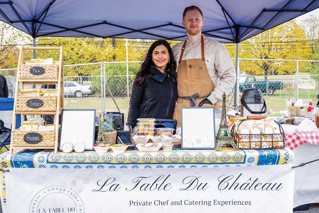 A man and woman standing under a tent, with a table in front covered in food items and displaying the sign "La Table du Chateau"