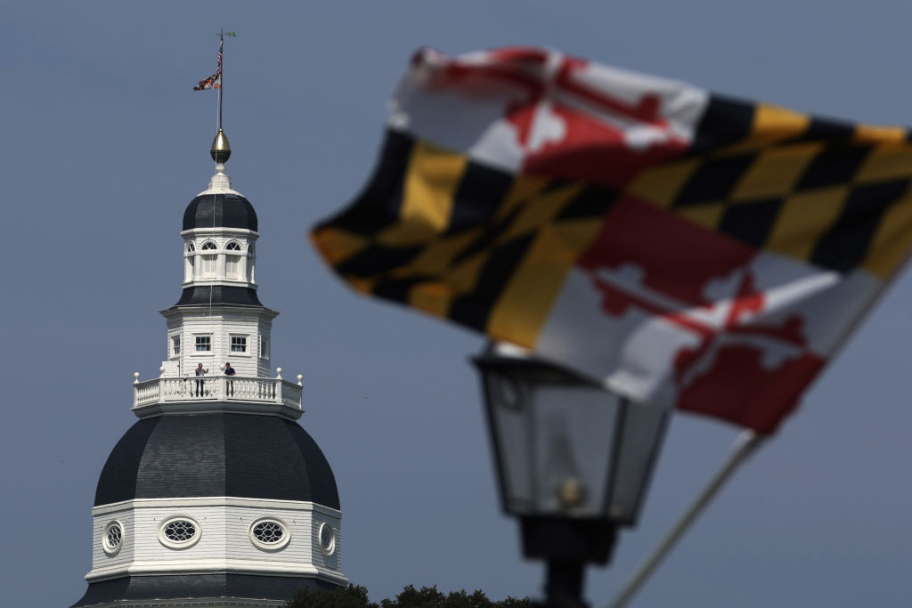 The flag of Maryland, red and white and black and yellow, in the foreground, the dome and tower of the Maryland State House behind.