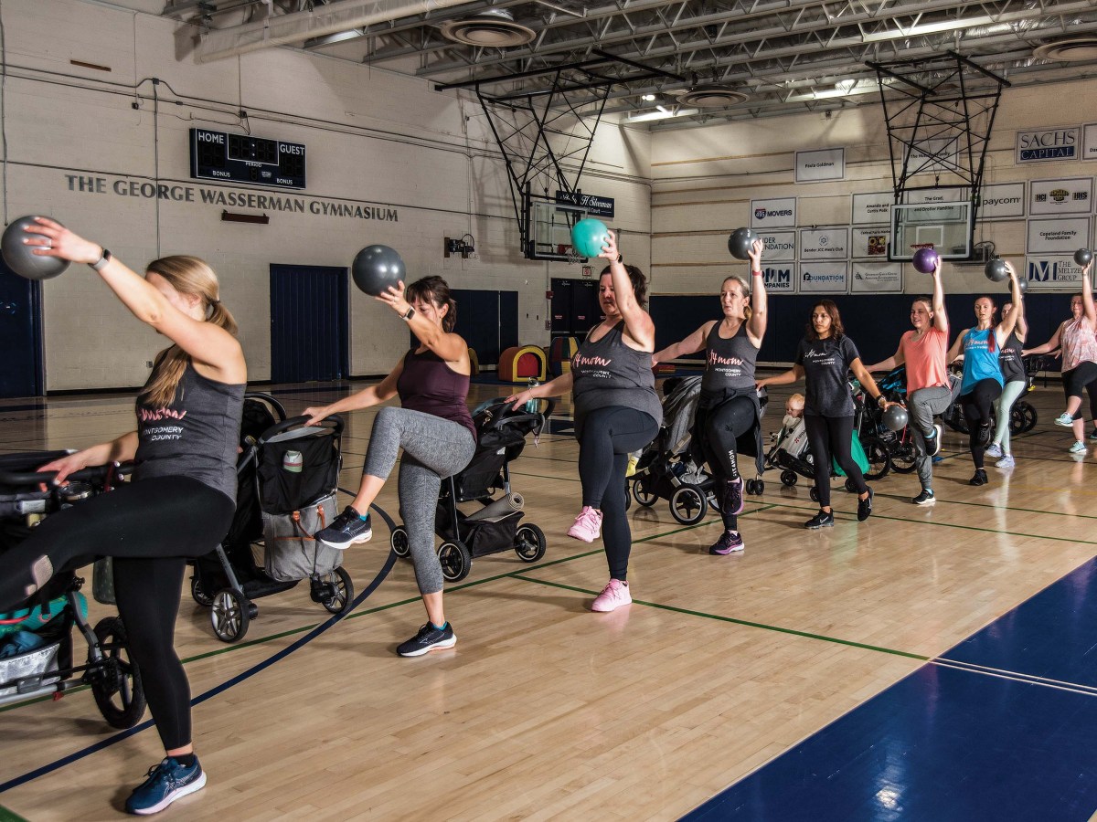 Women hold strollers in their right hand and lift exercise balls with their left inside a gymnasium